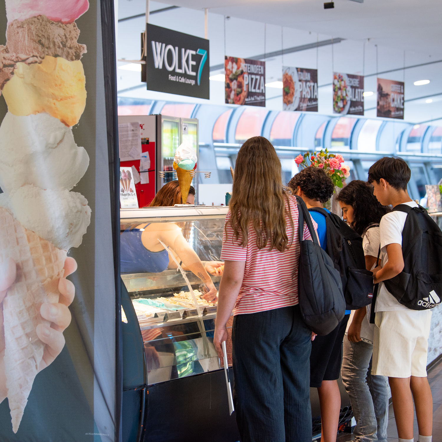 Mehrere Jugendliche stehen in einer Reihe an einer Eistheke im LuisenForum. Eine Verkäuferin reicht Eis über die Theke, im Hintergrund ist die Gastronomiezone „Wolke 7“ zu sehen.
