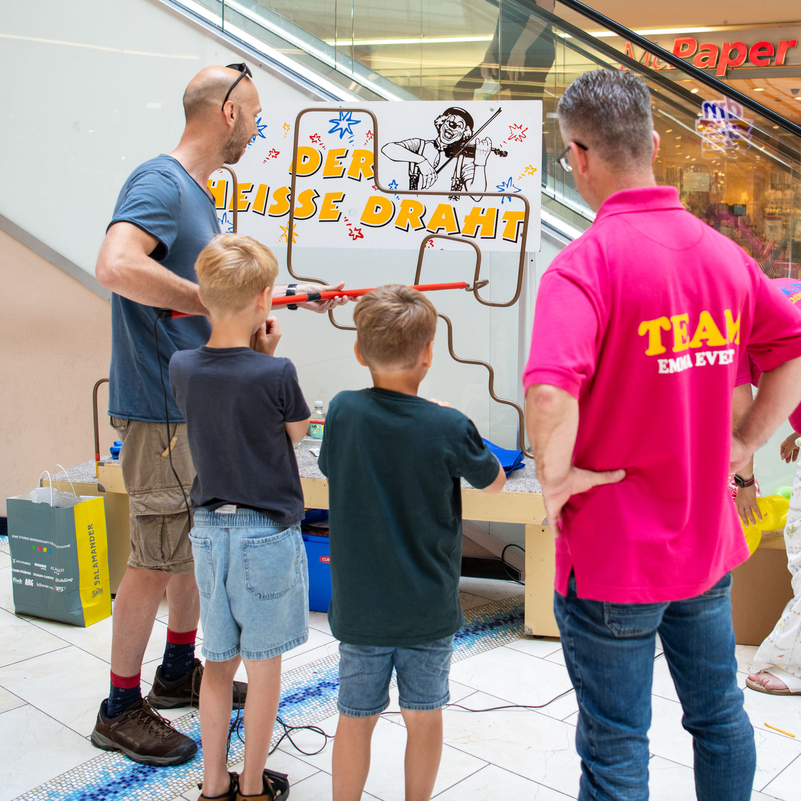 Ein Vater und zwei Jungen stehen vor dem Spiel „Der heiße Draht“, ein Event-Mitarbeiter in pinkem Shirt schaut zu.