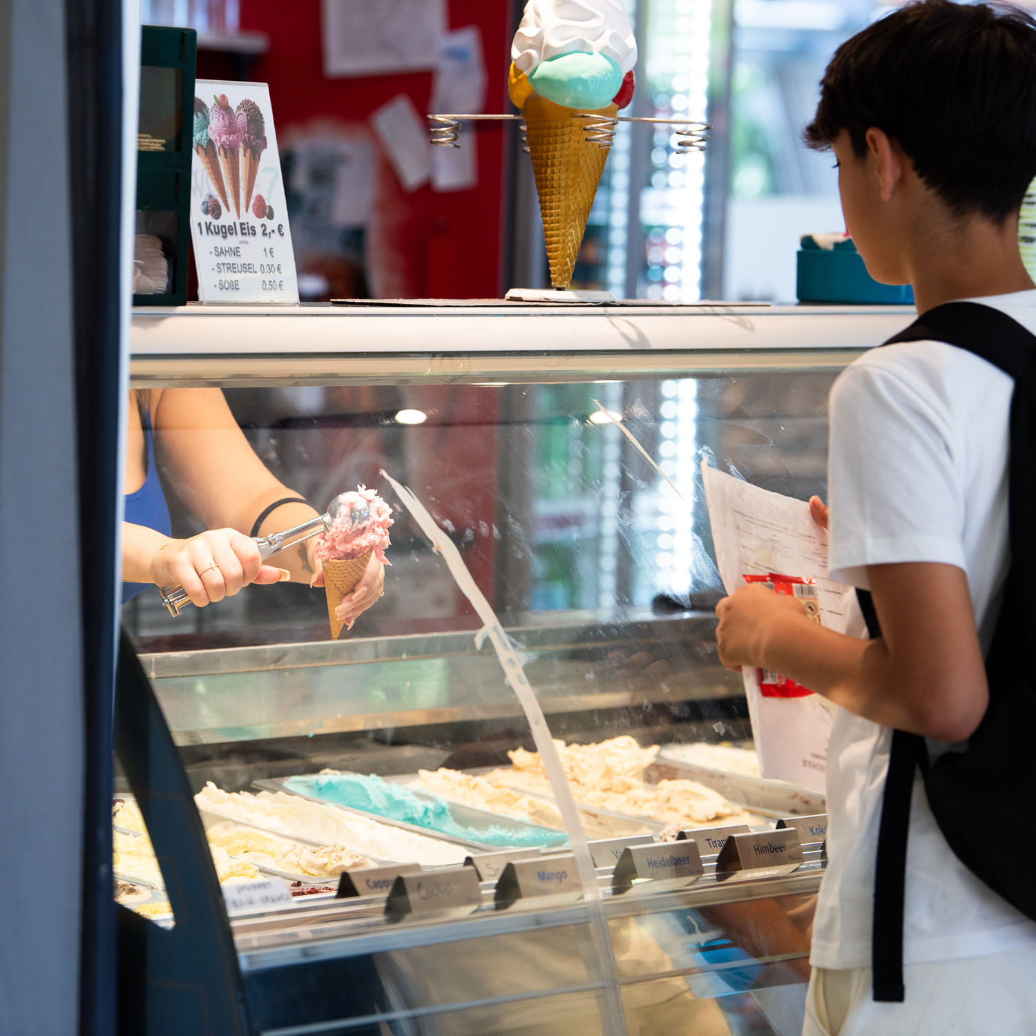 Ein Junge in weißem T-Shirt steht vor einer Eistheke und beobachtet, wie eine Verkäuferin eine rosa Eiskugel in eine Waffel portioniert. Auf der Theke steht ein Preisschild.