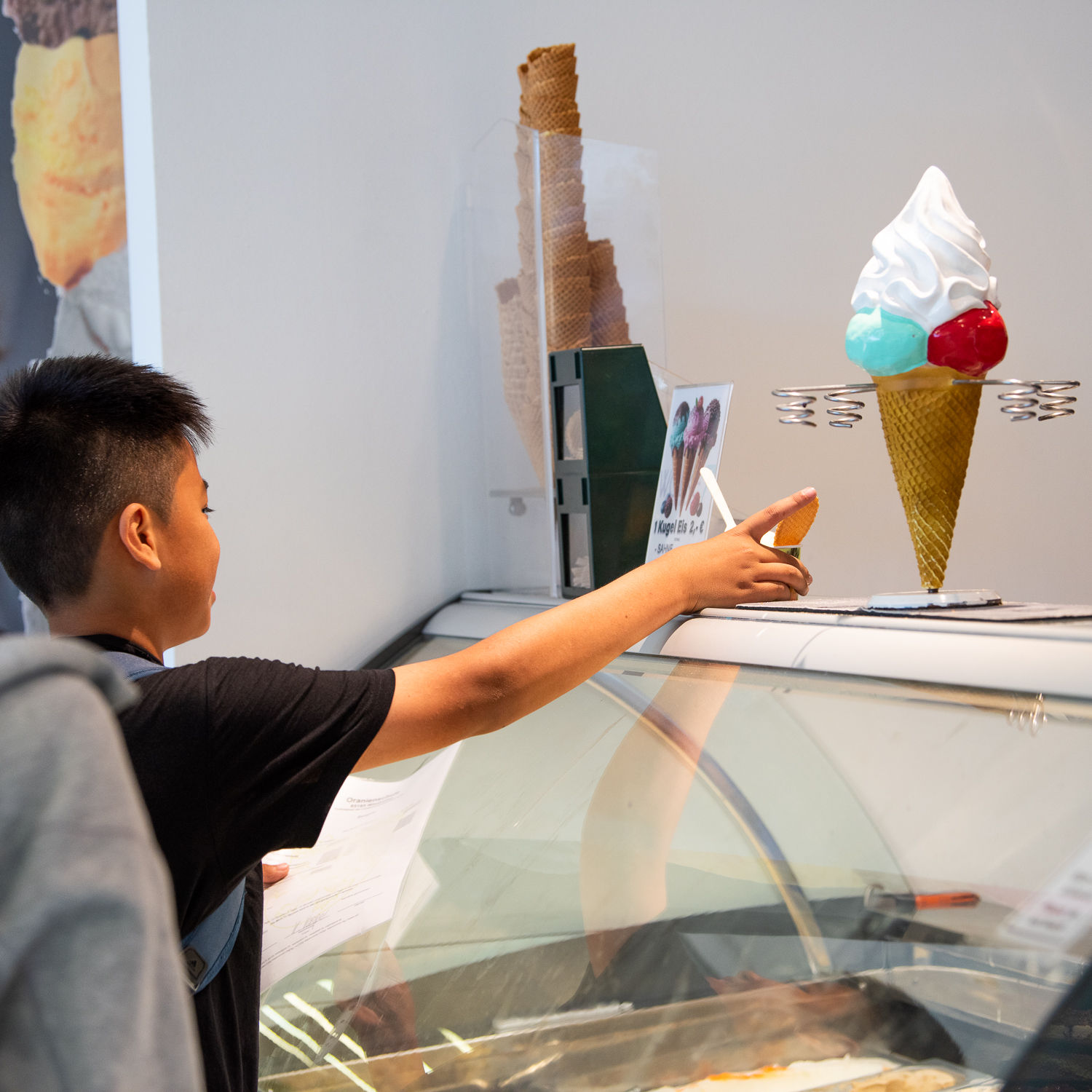 Ein Junge mit dunklen Haaren steht vor einer Eistheke und erhält eine Eiswaffel. Im Hintergrund sind Eiswaffeln, eine dekorative Eiskugel aus Kunststoff und ein Schild mit Preisen zu sehen.